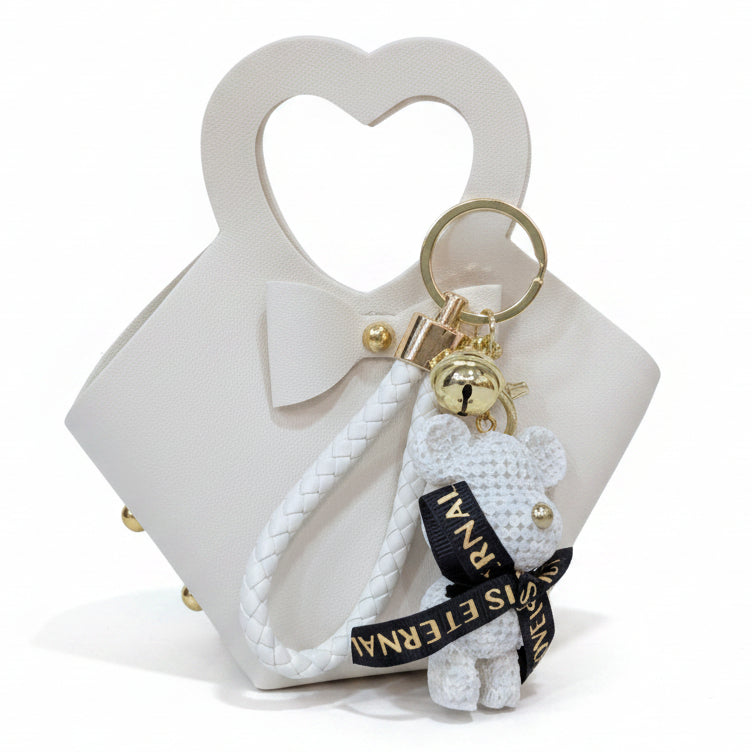 Borsetta bianca con manico a forma di cuore, fiocco frontale e portachiavi con orsetto bianco e nastro nero su sfondo bianco