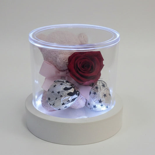 Cofanetto cilindico in acrilico trasparente su base luminosa bianca contenente un orsetto rosa, una rosa stabilizzata rossa e cioccolatini incartati su sfondo bianco